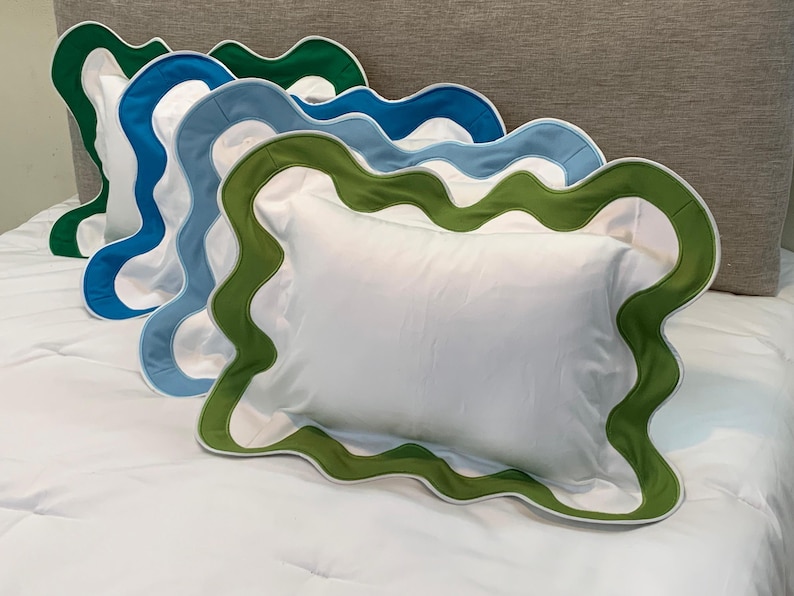 May include: Four white rectangular pillows with scalloped edges in green, blue, and white are arranged on a white bed. The pillows have a solid white center and a contrasting border. The pillows are placed on a white bedspread.
