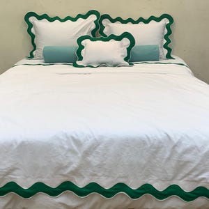 May include: White bed linens with green scalloped trim. The set includes a duvet cover, pillowcases, and decorative pillows. Two teal bolster pillows add a pop of color. The bedding is neatly arranged on a bed.