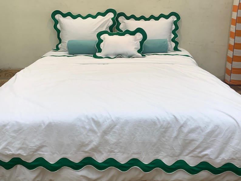 May include: White bed linens with green scalloped trim. The set includes a duvet cover, pillowcases, and decorative pillows. Two teal bolster pillows add a pop of color. The bedding is neatly arranged on a bed.