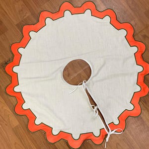 May include: A round, off-white Christmas tree skirt with a scalloped orange border and green trim. The skirt has a slit with ties for wrapping around the tree trunk. The design is simple and festive.