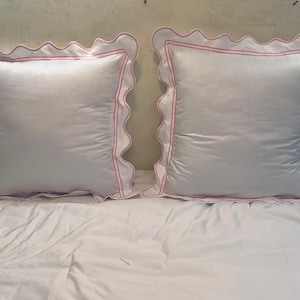 Può includere: Due cuscini quadrati bianchi con bordi rosa e bordi smerlati. I cuscini sono su un copriletto bianco. I cuscini hanno una lucentezza satinata.