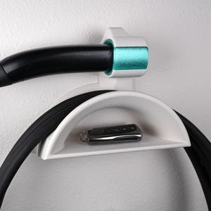 Puede incluir: Un soporte de pared blanco para cable de carga de vehículo eléctrico con un estante curvo para almacenar el cable y una base superior para el mango de carga. El mango es negro con un acento turquesa. Una pequeña llave de coche negra descansa sobre el estante.