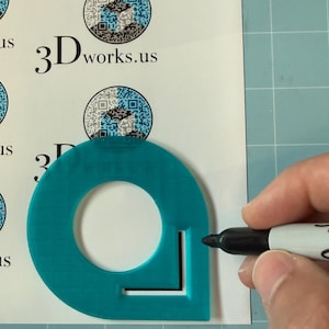 May include: A teal plastic corner rounding template with a circular cutout and a black marker positioned on a white sheet featuring multiple printed circular logos with the text 3Dworks.us and a QR code, set against a green cutting mat.