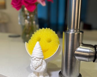 Op de afbeelding: Een witte sponshouder in de vorm van een kabouter met een getextureerde baard en een puntmuts, die een ronde gele schuurspons met een lachend gezicht vasthoudt. De houder staat naast een roestvrijstalen keukenkraan.