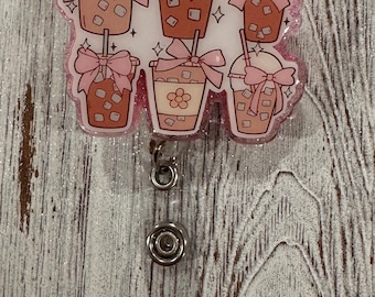 May include: A pink glitter badge reel with a charm featuring six iced coffee cups with pink bows and straws.