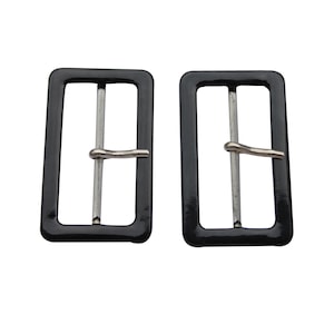 May include: Two black rectangular belt buckles with a silver bar and pin. The buckles have a glossy finish and are set against a white background. These buckles are suitable for belts, bags, or other accessories.