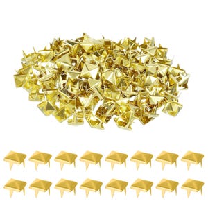 Può includere: Una collezione di borchie dorate a forma di piramide. Le borchie hanno una base quadrata e una parte superiore a punta. Un gruppo più piccolo di borchie è disposto sotto il mucchio più grande, mostrando il perno sul retro. Queste borchie decorative sono adatte a vari progetti artigianali.