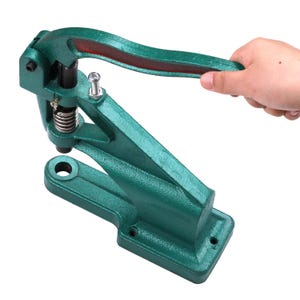 May include: A green metal grommet press with a red accent on the handle. The press has a spring and a lever for operation. The base has a rectangular shape with a hole for mounting.