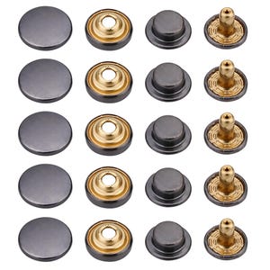 Può includere: Una collezione di sedici bottoni e rivetti per jeans in metallo grigio scuro. I bottoni sono rotondi e piatti, mentre i rivetti hanno un perno dorato e un cappuccio grigio scuro. Gli articoli sono disposti in quattro file da quattro.