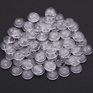 May include: A large collection of round, clear plastic buttons scattered across a dark surface. Each button features a smooth, transparent finish and two small holes in the centre for attachment, creating a uniform and functional sewing supply set.