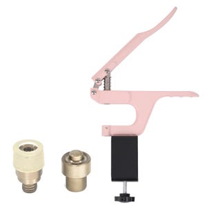 Pode incluir: Uma ferramenta de prensagem manual de metal rosa com uma base preta e dois acessórios de metal. A ferramenta é usada para colocar ilhós e botões de pressão.