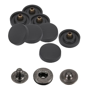 May include: A collection of black snap fasteners with brass-colored centers. The image shows several assembled snaps and individual components, including decorative metal snap heads with various designs. These fasteners are likely for clothing or crafts.