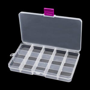 May include: A rectangular, clear plastic storage container featuring fifteen individual compartments arranged in a grid. The lid is attached with a hinge and includes a bright magenta latch mechanism at the top centre for secure closure.