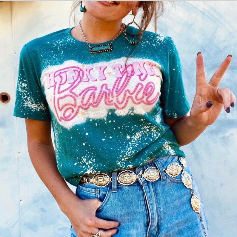 May include: A teal blue t-shirt with a bleach-splattered design. The shirt features a pink and white graphic that says "Howdy Y'all Barbie". The person wearing the shirt is also wearing a silver belt with decorative medallions.