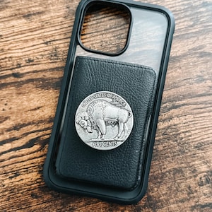 May include: A black phone case with a clear back and a black leather wallet attached to the back. The wallet has a silver buffalo nickel coin attached to it. The coin reads "United States of America" and "Five Cents".