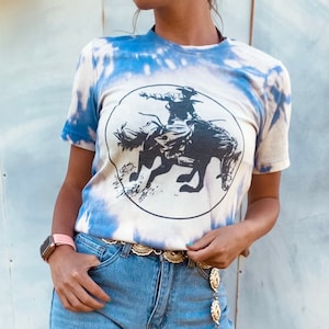 K&ouml;nnte beinhalten: Ein wei&szlig;es T-Shirt mit einem blauen Batik-Muster. Das Shirt zeigt eine schwarz-wei&szlig;e Grafik eines Cowboys, der ein Pferd reitet. Das Shirt hat einen runden Ausschnitt und kurze &Auml;rmel.