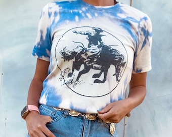 May include: A white t-shirt with a blue tie-dye pattern. The shirt features a black and white graphic of a cowboy riding a horse. The shirt has a round neckline and short sleeves.