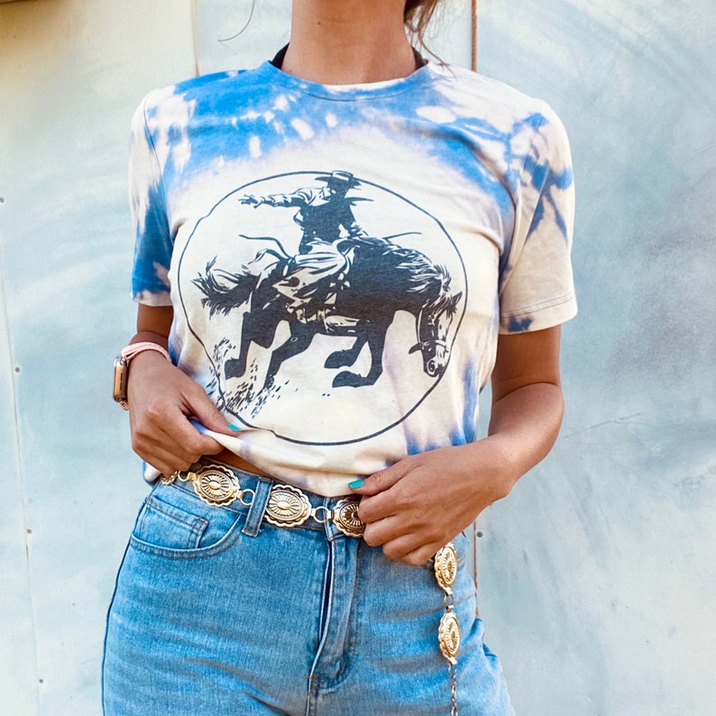 K&ouml;nnte beinhalten: Ein wei&szlig;es Batik-T-Shirt mit einem blauen und wei&szlig;en Cowboy-Motiv, der ein Pferd reitet. Das Shirt hat einen runden Ausschnitt und kurze &Auml;rmel. Das Model tr&auml;gt einen goldenen Ketteng&uuml;rtel mit dekorativen Medaillons.
