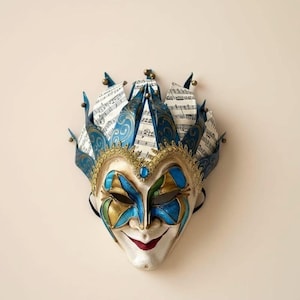 May include: A jester mask featuring a cream-colored face with red lips, blue and green eye accents, and a gold-trimmed forehead. The crown consists of blue patterned fabric panels interspersed with white paper printed with black musical notes.