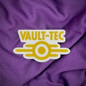 May include: A white sticker with a yellow Vault-Tec logo. The logo features the Vault-Tec text and a stylized symbol with three white lines and a yellow circle.