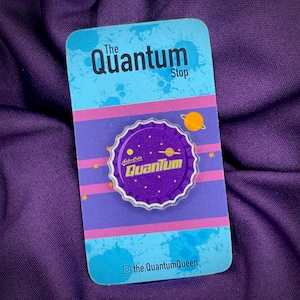 May include: Purple and gold Quantum Cola bottle cap pin with a white border and stars. The pin is on a blue and pink striped card with the text "The Quantum Stop" and "@the.quantumQueen".
