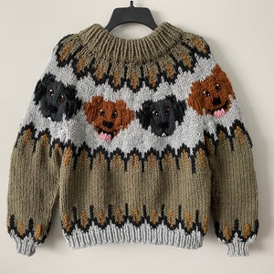 Puede incluir: Un suéter de punto verde, gris y marrón con un patrón de punto de trenza y tres caras de perro. Las caras de perro son marrones, negras y negras y marrones.