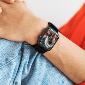 May include: Black smartwatch with a black strap. The watch face shows a colourful image of a white creature with a floral background. The watch is worn on a wrist, with a denim shirt and red shirt in the background.