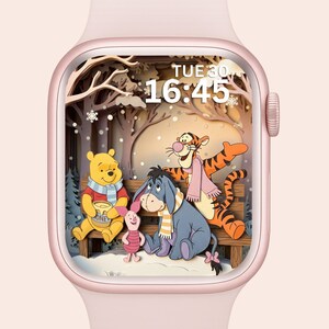 Op de afbeelding: Een roségouden Apple Watch toont een winterse scène met Winnie de Poeh, Knorretje, Iejoor en Tijger. De wijzerplaat toont de tijd als 16:45 uur en de dag als dinsdag. De personages zitten op een houten bank in een besneeuwd bos.