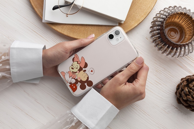 Op de afbeelding: Een transparante telefoonhoes met een bedrukte afbeelding van Mickey Mouse en Minnie Mouse die elkaar kussen. De hoes is ontworpen voor een smartphone met een dubbele camera lens.
