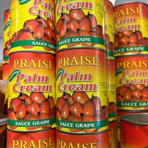 Op de afbeelding: Een stapel rode en gele blikken Praise Palm Cream Sauce Graine. De blikken zijn gelabeld met de merknaam, productnaam en ingrediënten. De blikken zijn gestapeld in een supermarkt.