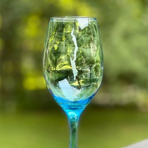 May include: A blue glass wine glass with a white etched design of a lake or island. The glass has a clear stem and a blue base.