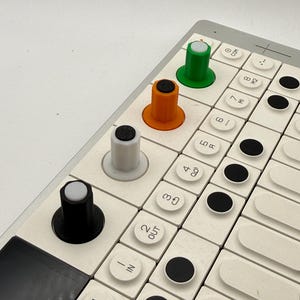 Könnte beinhalten: Ein weißes Interface für einen elektronischen Musik-Synthesizer mit einem Raster aus quadratischen Tasten mit schwarzen kreisförmigen Akzenten und vier hohen, zylindrischen Drehreglern in Schwarz, Weiß, Orange und Grün. Die Tasten sind beschriftet.