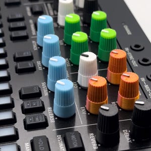Könnte beinhalten: Nahaufnahme eines schwarzen Musik-Synthesizers mit verschiedenen Knöpfen und Tasten. Die Knöpfe sind in den Farben Blau, Grün, Weiß und Orange. Die Tasten sind mit "Delay", "Reverb", "Env", "LFO", "Noise", "Attack", "Decay", "Sustain", "Release", "Destination", "D-Motion", "Hold", "Manual", "Porta On", "Porta Time" und "Poly" beschriftet.