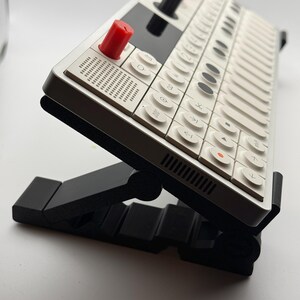 Könnte beinhalten: Ein tragbarer Synthesizer mit weißem Gehäuse und einem Raster aus weißen Tasten, ausgestattet mit einem markanten roten Drehknopf, einem schwarzen Display sowie weiteren grünen und orangen Bedienelementen auf einem verstellbaren schwarzen Ständer.