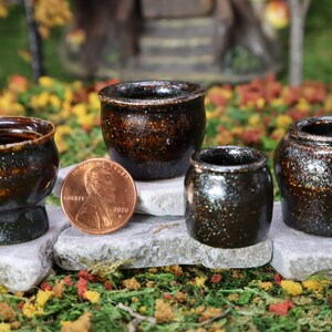 May include: Four miniature brown and black speckled ceramic pots with a shiny glaze. The pots are arranged on small gray stones. A US penny is placed next to one of the pots for scale.