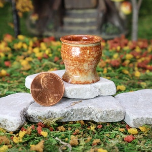 May include: A small, brown and white ceramic pot with a flared rim, sitting on top of three gray stones. A penny is placed next to the pot for scale.