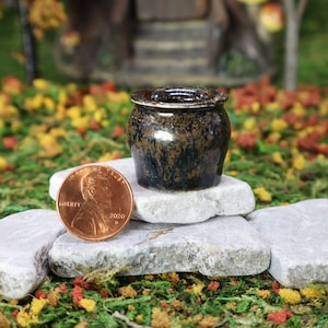 May include: A small, dark brown ceramic pot with a black glaze sits on a stack of three white stones. A penny is in the foreground for scale.