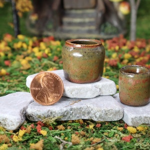 May include: Two small, speckled, brown ceramic pots with a green glaze, sitting on top of gray rocks. A penny is placed on the rock below the larger pot.