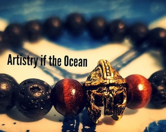 May include: A black lava rock and red tiger's eye beaded bracelet with a gold gladiator helmet charm. The bracelet has the text "Artistry if the Ocean" on it.