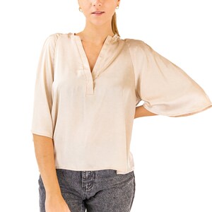 May include: A woman wearing a light beige blouse with a v-neck and a button closure. She is also wearing dark gray jeans.