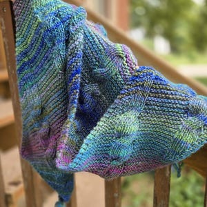 May include: A hand-knitted triangular shawl featuring a variegated yarn pattern in shades of blue, green, and purple. The fabric displays a textured cable knit design throughout the body and includes a small tassel at the pointed end of the garment.