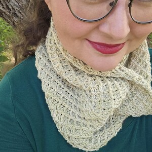 May include: A light beige crocheted cowl scarf with a textured pattern. The scarf is wrapped around the neck and shoulders. The person is wearing a dark green shirt and glasses.