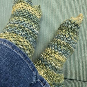 May include: A pair of hand-knitted socks in shades of green, blue, and yellow. The socks are thick and textured, with a ribbed cuff and a pointed toe. The socks are worn with blue jeans.