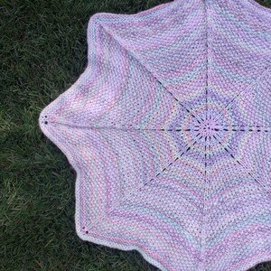 May include: A star-shaped crocheted baby blanket in shades of pink, purple, and blue. The blanket is made with a soft yarn and has a delicate, lacy pattern.