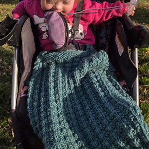 May include: A teal knitted blanket with a textured pattern is draped over a stroller. The blanket is made of a soft yarn and has a ribbed design. The stroller is black and has a child in a pink jacket.
