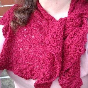 May include: A crimson red crochet shawl with an intricate openwork design. The shawl has a scalloped edge and a draped style. The soft texture and rich colour make it a versatile accessory. The shawl is handmade, adding a touch of elegance to any outfit.