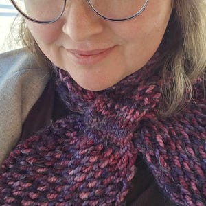 May include: A close-up of a person wearing a purple and pink knitted scarf. The scarf is made of a chunky yarn and has a textured look. The person is also wearing a gray hat and glasses.