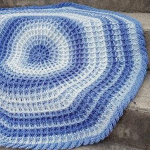 May include: A round, crocheted blanket in shades of blue and white. The blanket features a waffle stitch pattern that creates a textured surface. The design spirals from the center outwards, with the colors gradually changing from light to dark blue.