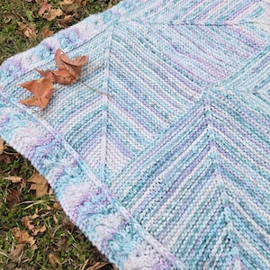 May include: A knitted baby blanket in shades of blue, purple, and white with a chevron pattern. The blanket has a decorative border.