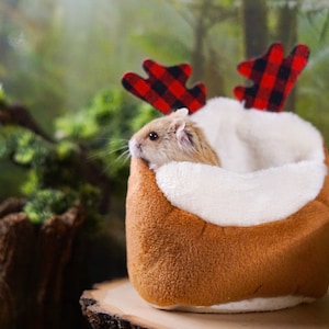May include: A small hamster wearing a red and black plaid reindeer antler headband sits in a brown and white plush bed.
